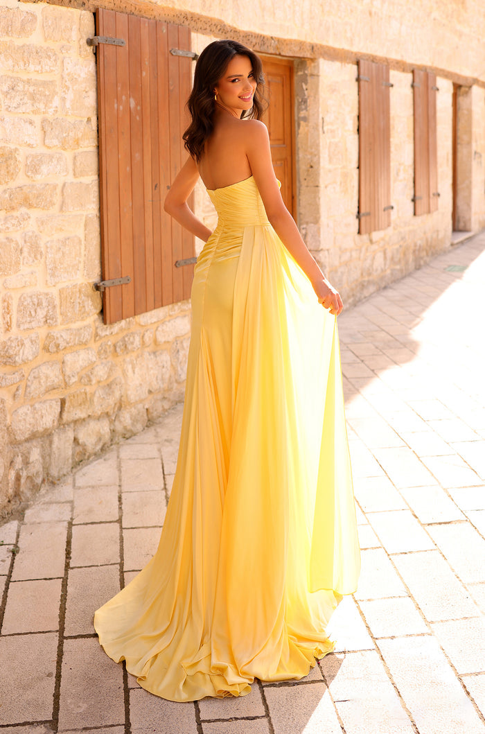 A woman in a flowing yellow strapless gown stands against a stone wall with wooden shutters.