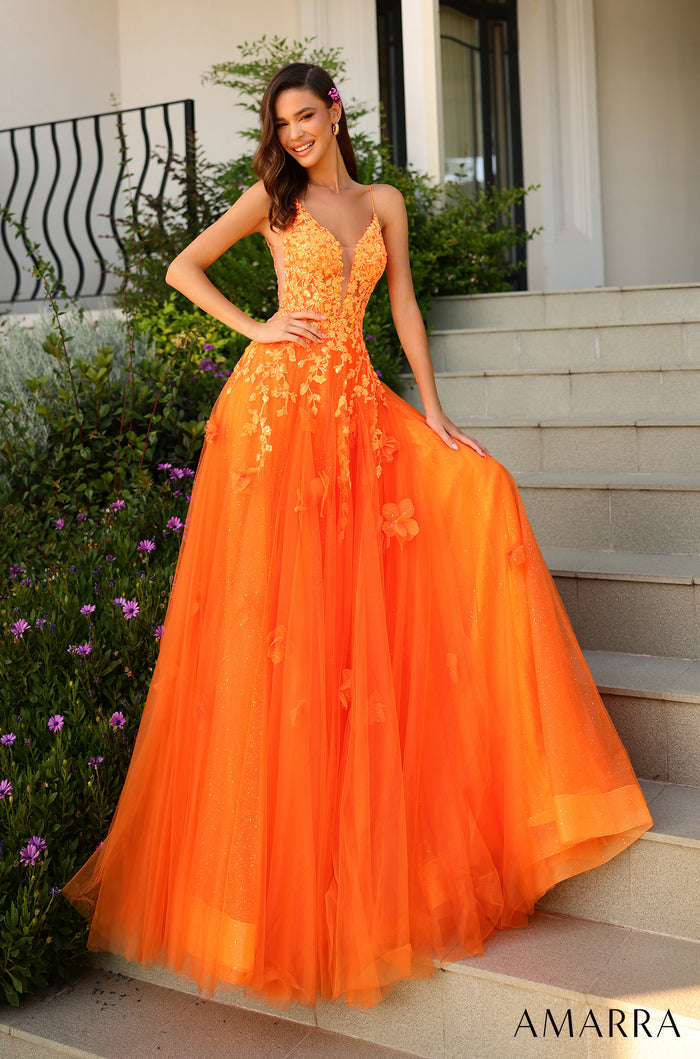 A smiling model wears a full, floor-length neon orange tulle gown with floral lace bodice on stone steps.
