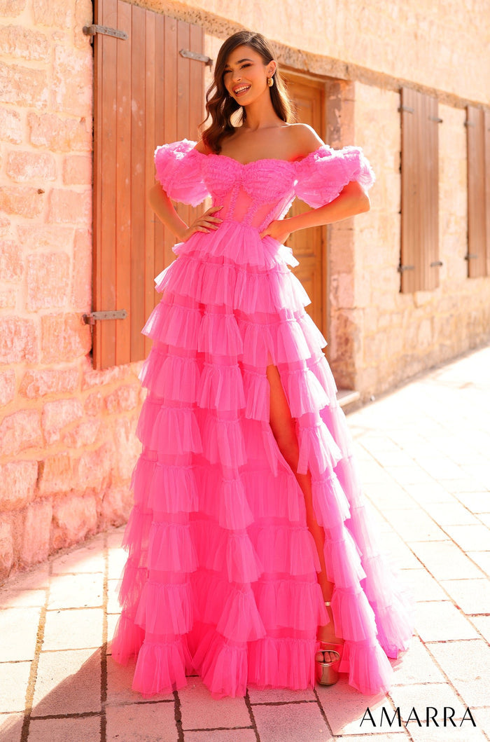 A smiling woman wears a vibrant pink tiered tulle gown with off-shoulder ruffles against a stone wall backdrop.