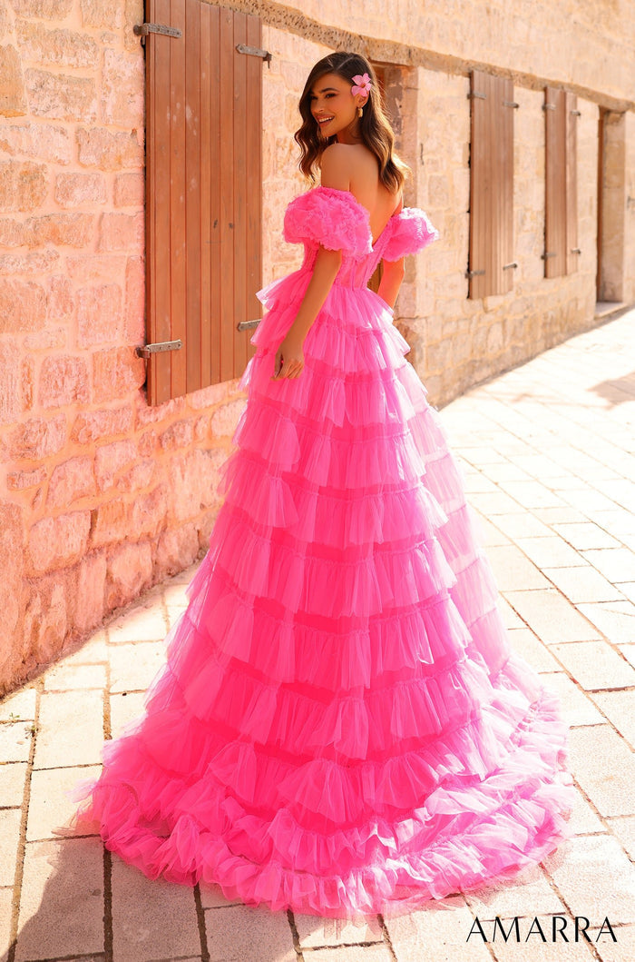 A person wears a vibrant pink tiered tulle ball gown with off-shoulder ruffles against a stone wall background.