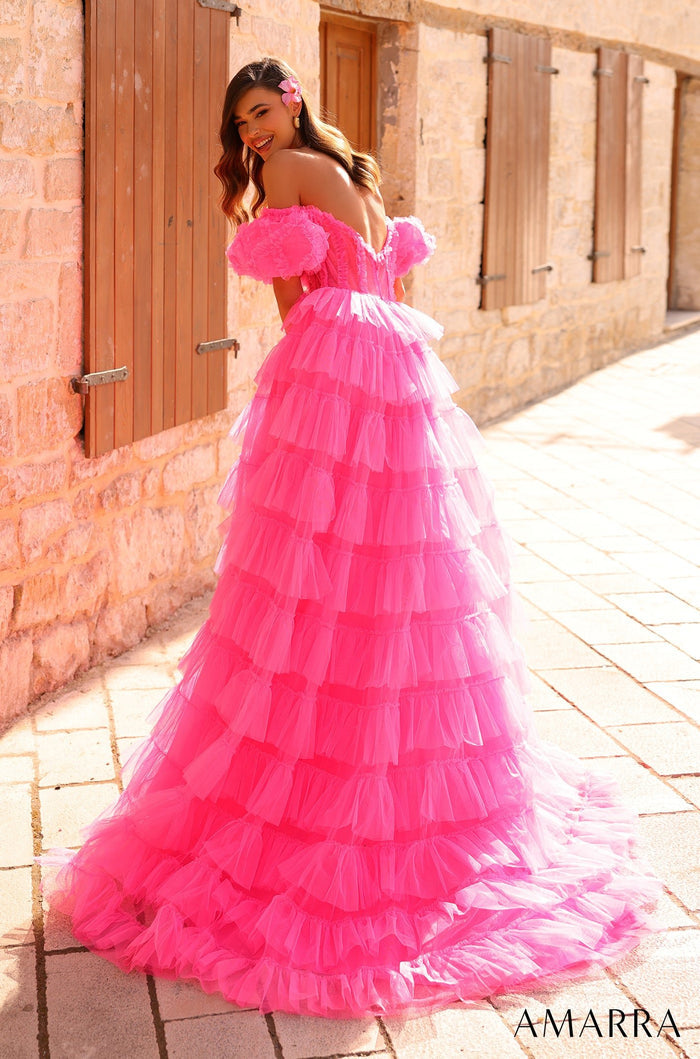 A woman wears a hot pink tiered tulle ball gown with off-shoulder ruffles, standing against a stone wall.