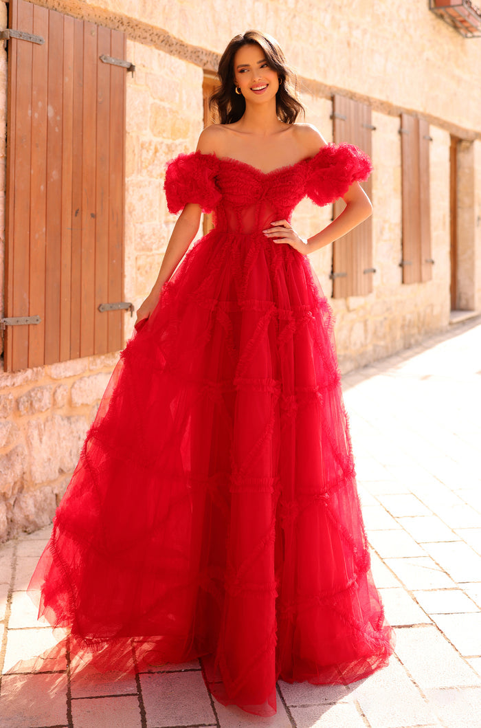 A woman wears a dramatic red tulle ball gown with off-shoulder ruffles, posing against a stone wall backdrop.
