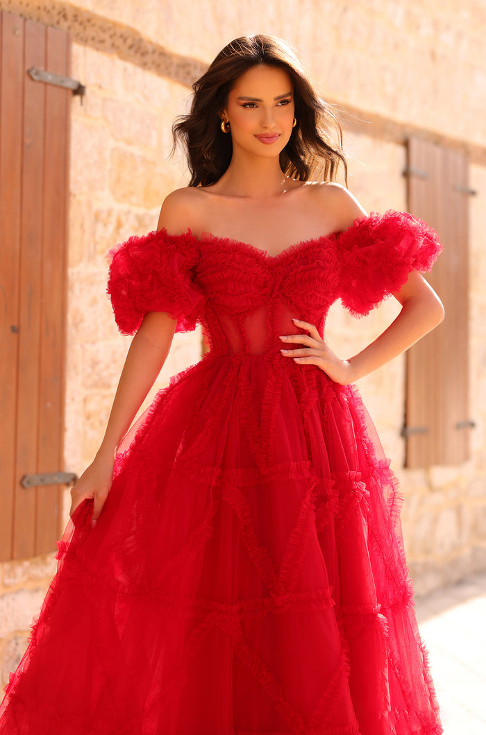 A woman wears a vibrant red, off-shoulder tulle ball gown with ruffled layers against a stone wall backdrop.