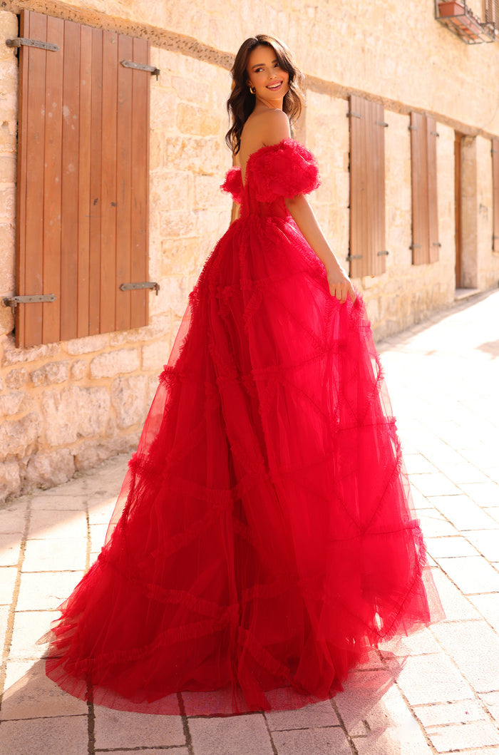 A woman in a dramatic, full-skirted red tulle gown poses against a stone wall with wooden shutters.