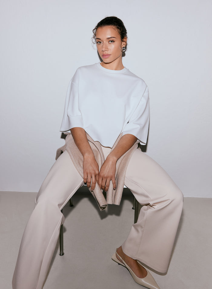 Nainen yllään valkoinen Luxe Boxy Short Sleeve -toppi, vaaleanbeiget leveälahkeiset housut ja vaaleanbeiget ballerinakengät, istumassa tuolilla, neule sylissään. [M-170cm]