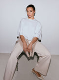 Nainen yllään valkoinen Luxe Boxy Short Sleeve -toppi, vaaleanbeiget leveälahkeiset housut ja vaaleanbeiget ballerinakengät, istumassa tuolilla, neule sylissään. [M-170cm]