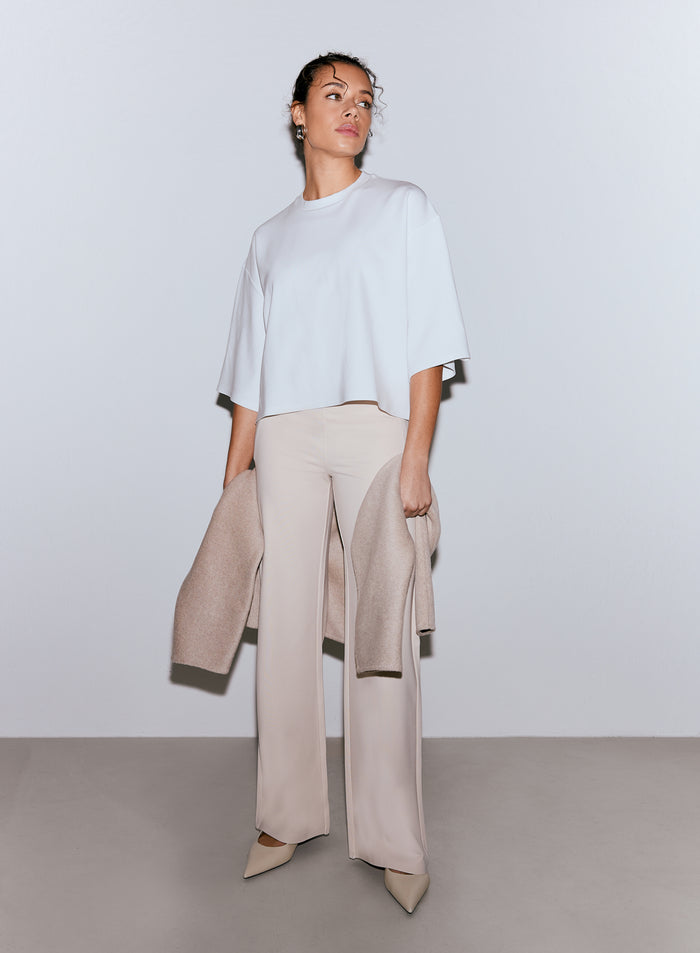 Malli yllään valkoinen Luxe Boxy Short Sleeve -toppi ja vaaleanbeiget leveälahkeiset housut, pidellen vaaleanbeigeä takkia. [M-170cm]